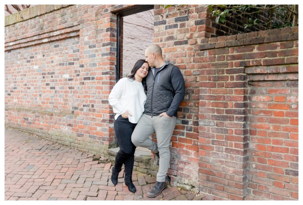 Winter Engagement Photos Alexandria VA Waterfront - man kisses woman's forehead in front of brick wall